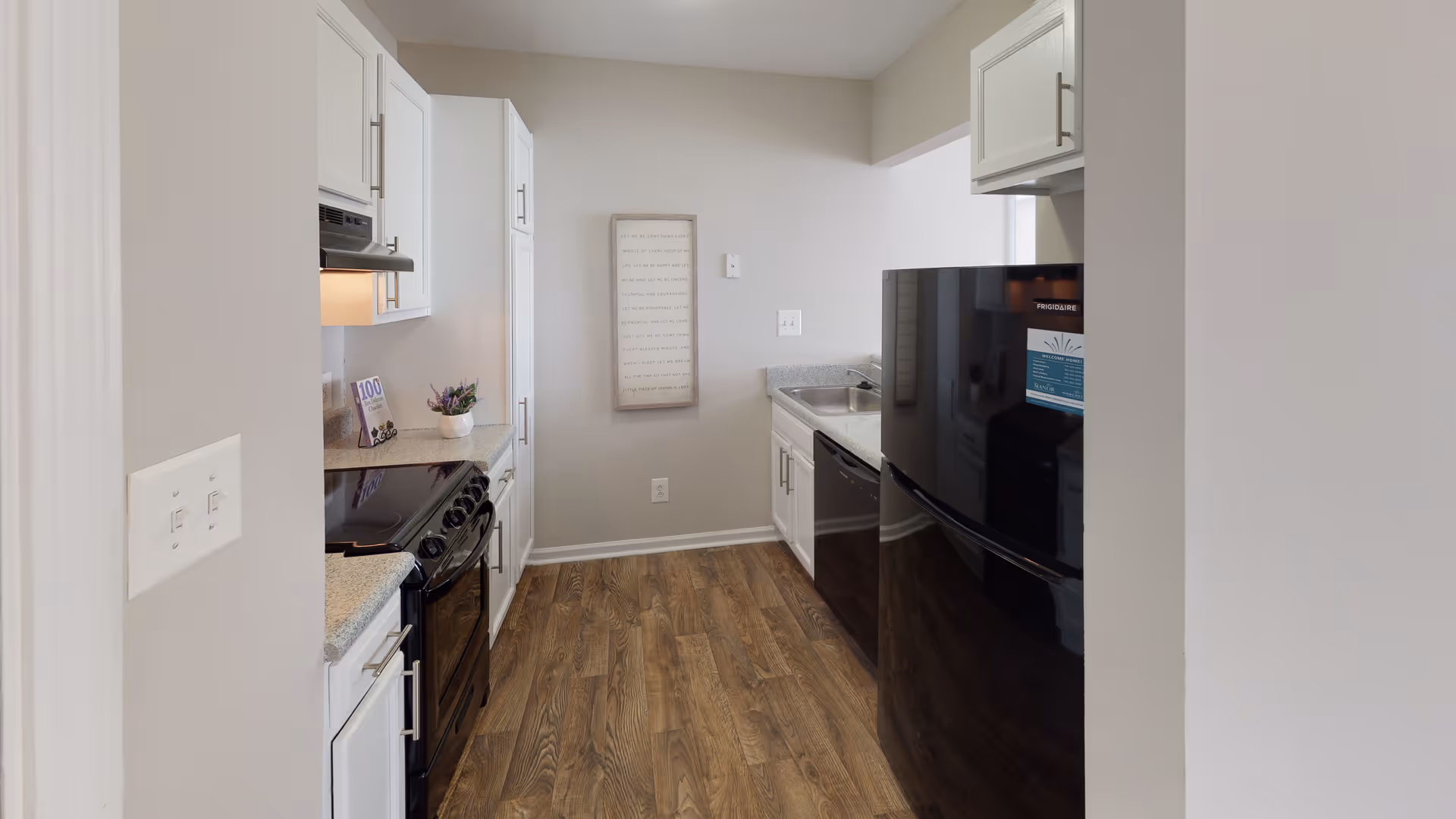 A modern kitchen with white cabinets, a black refrigerator, a black stove with an overhead vent, a dishwasher, and a countertop with a small plant and a decorative item. The floor is wood, and there is a framed piece of art on the far wall.