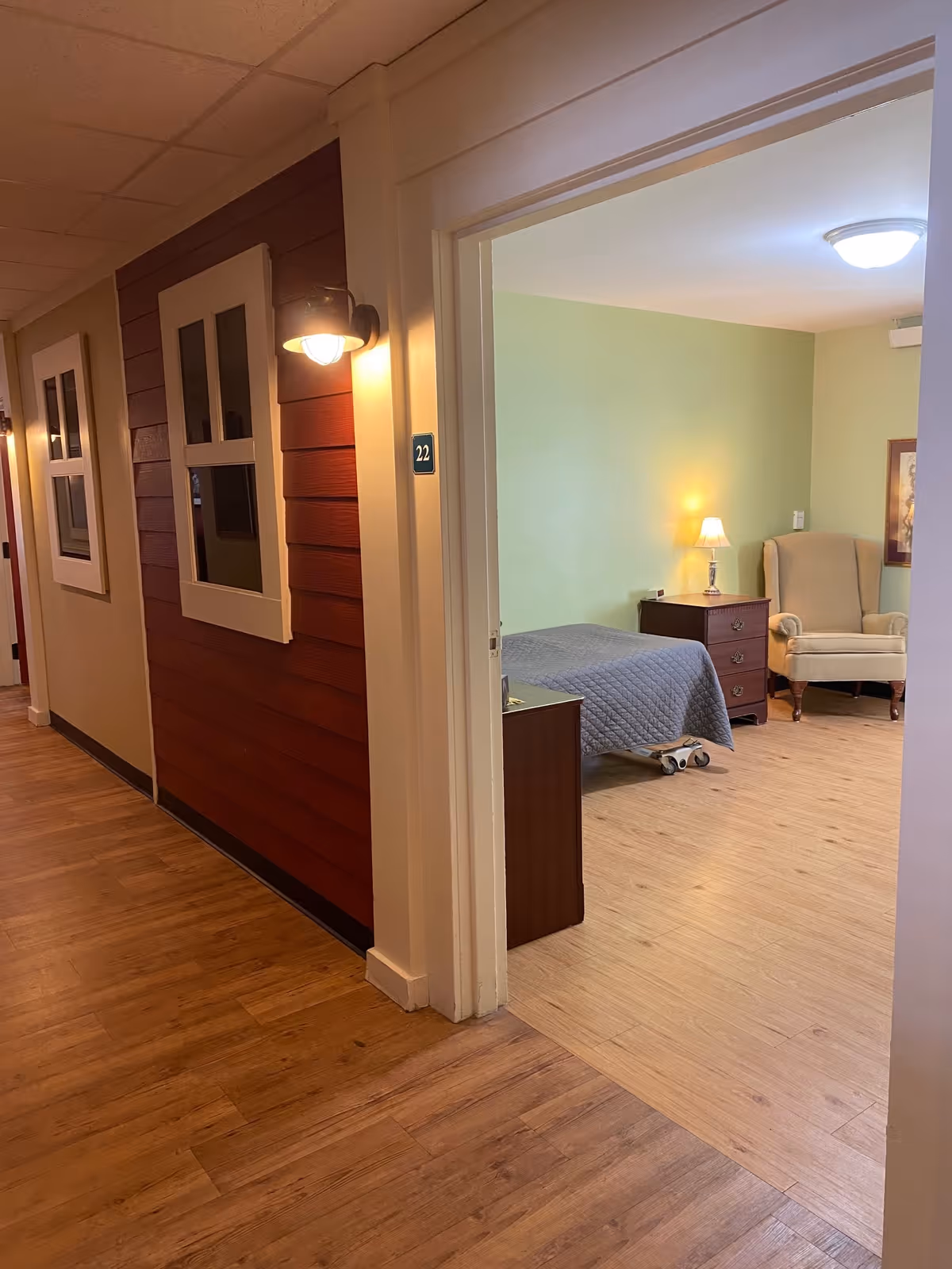 View down a hallway with wooden flooring and walls, leading to an open doorway of a room numbered 22. Inside the room, there is a bed with a gray quilt, a wooden nightstand with a lamp, and a beige upholstered armchair against a light green wall.
