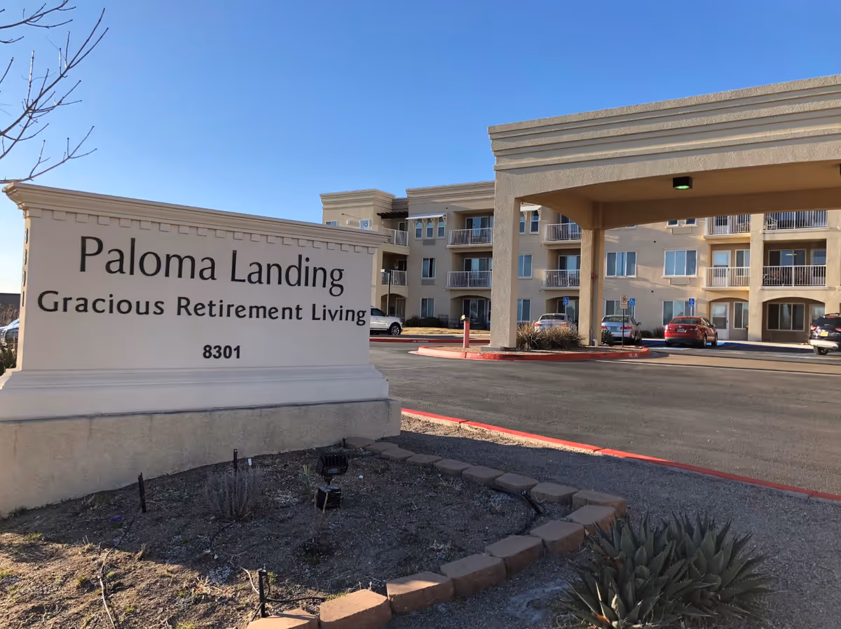Front entrance sign reading 'Paloma Landing Gracious Retirement Living' with the facility building, porte-cochère, and parking area visible behind it.