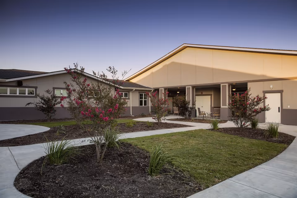 Exterior front entrance of a single-story memory care building with curved walkways, landscaped beds with flowering shrubs, and a covered porch with seating.