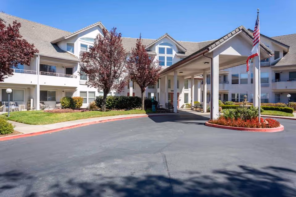 Exterior view of a senior living facility with a covered entrance, two trees with red leaves, well-maintained landscaping, and an American flag on a flagpole.