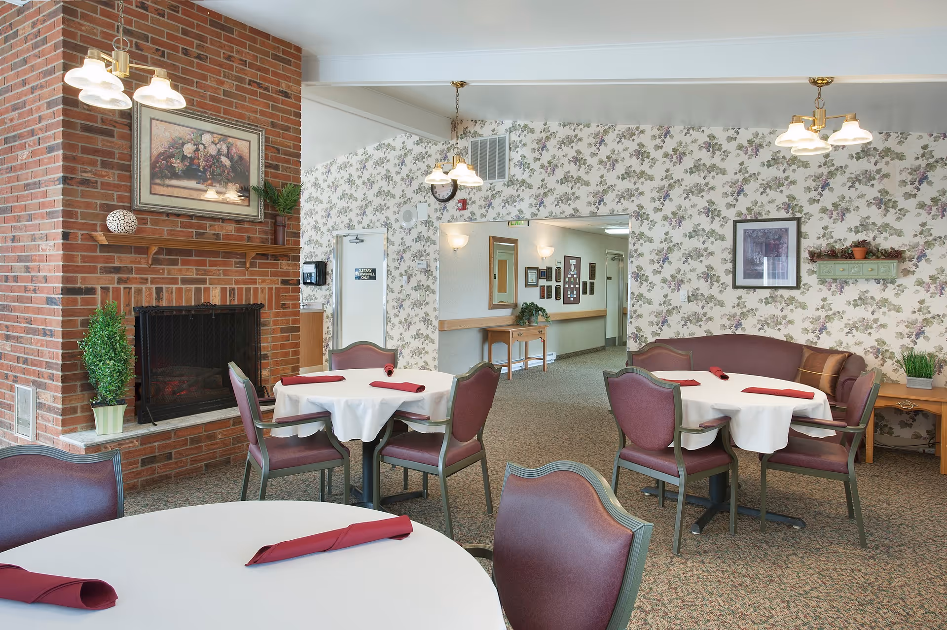 A cozy dining area in Albany Health and Rehabilitation Center featuring round tables covered with white tablecloths and burgundy napkins. The room has floral wallpaper, a brick fireplace with a framed floral painting above it, and several chairs around the tables. There are hanging light fixtures and some decorative plants placed around the room.
