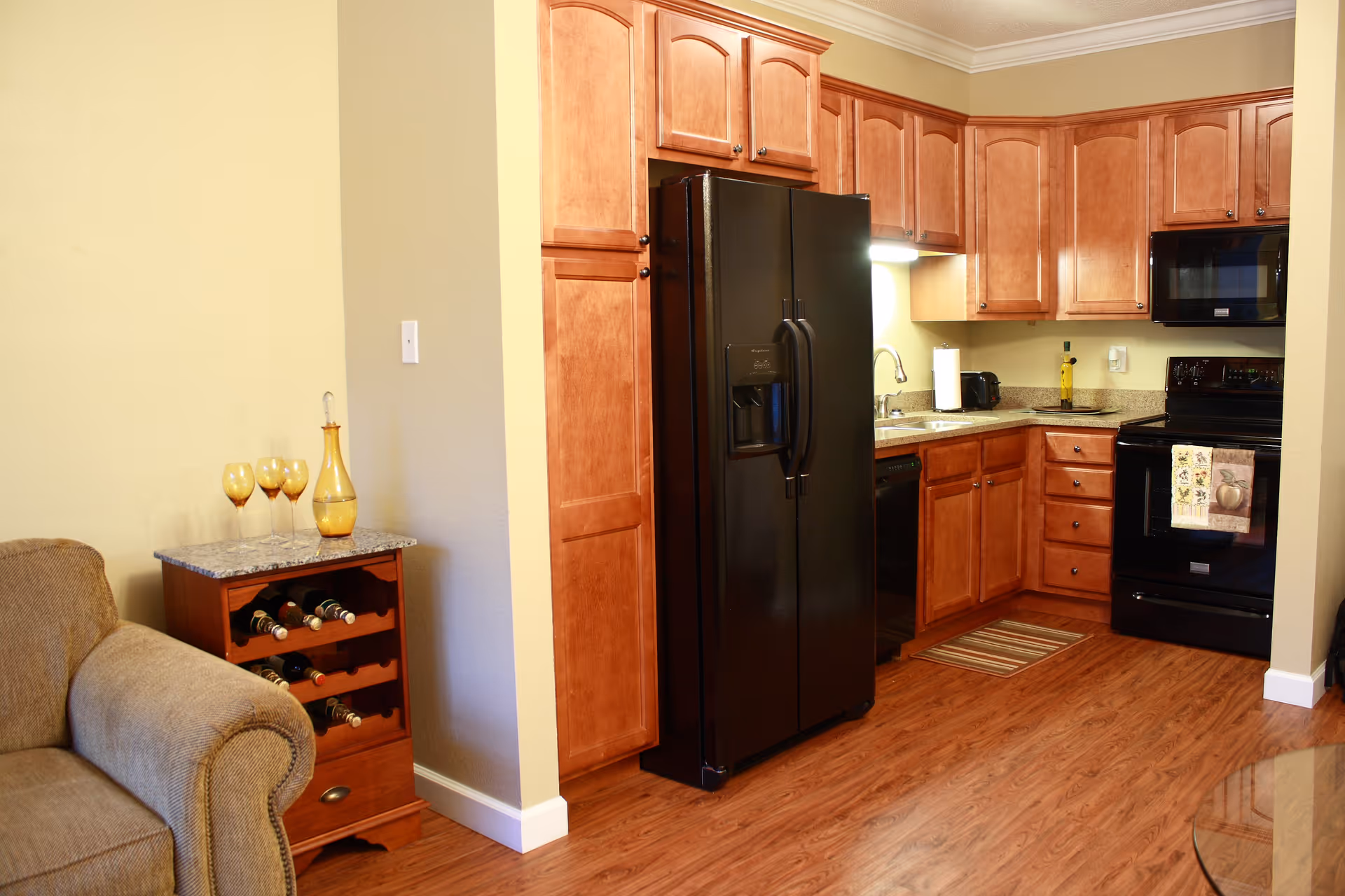 A cozy kitchen area with wooden cabinets, a black refrigerator, black stove, and microwave. The kitchen has a granite countertop with a sink and various small appliances. To the left, there is a small wooden wine rack with several bottles and three amber-colored glasses on top. Part of a beige upholstered armchair is visible in the foreground.