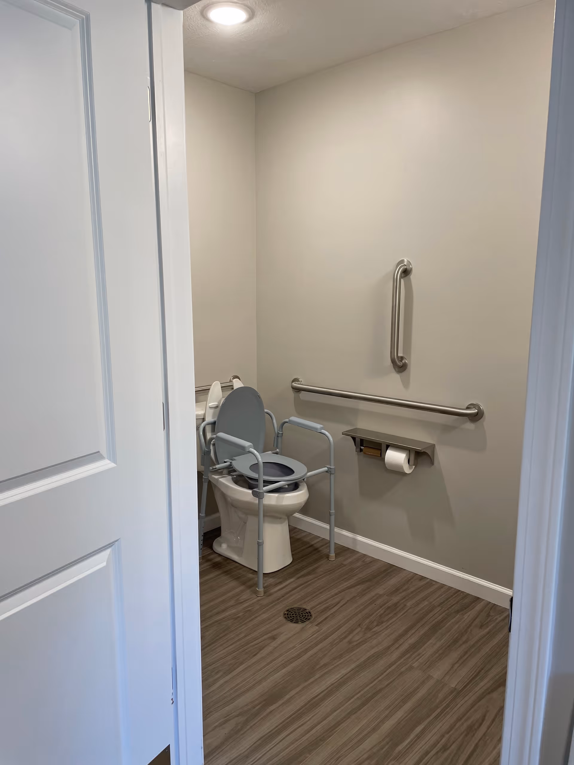 View into a bathroom with a toilet equipped with a raised seat and arm supports for accessibility. The bathroom has grab bars on the wall, a toilet paper holder, and wood-look flooring. The walls are painted light beige and the door is partially open.