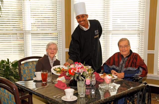Two elderly residents seated at a dining table smiling as a chef serves their meals.