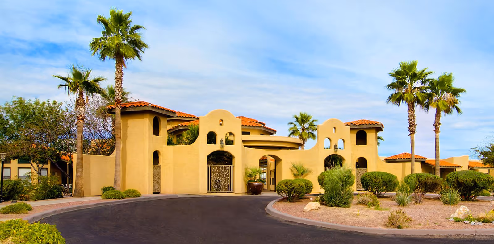 Front entrance of a Southwestern-style senior living building with stucco walls, terracotta roofs, palm trees, and a circular driveway.