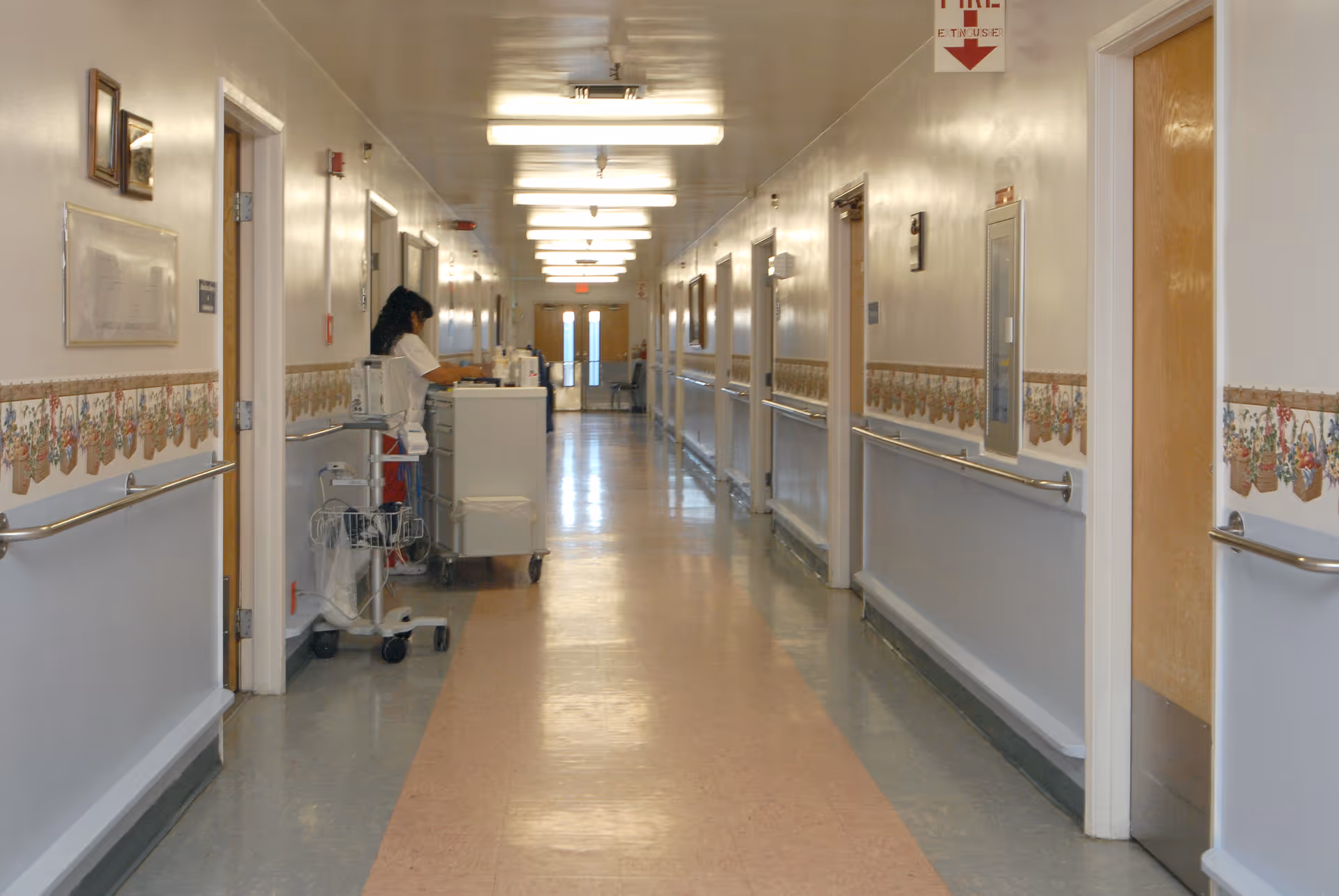 Long interior hallway of a convalescent hospital with handrails, doors along both sides, fluorescent lights, and a staff member at a medical cart.