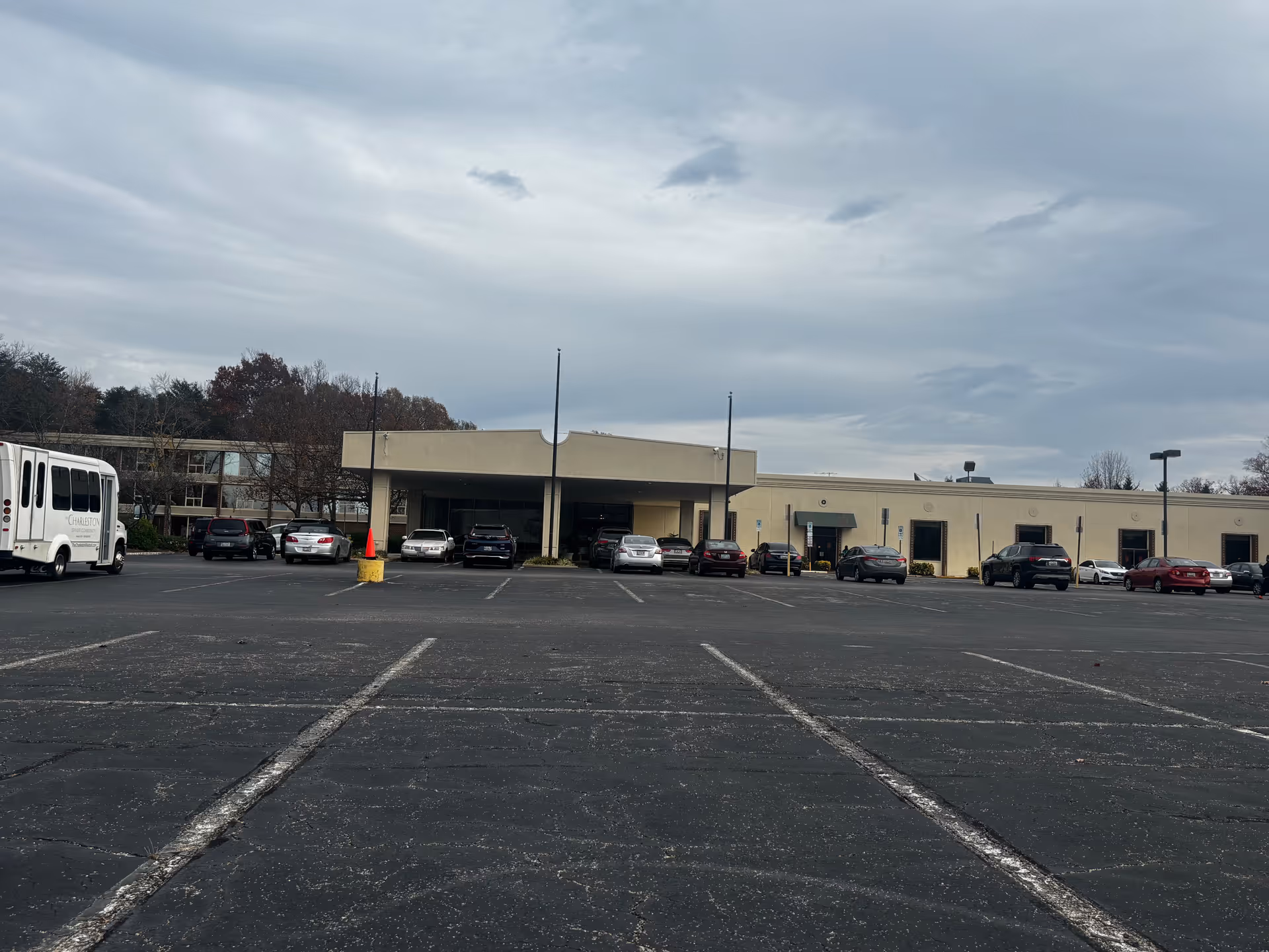 Exterior view of The Charleston Senior Community building with a large parking lot in the foreground. Several cars are parked near the entrance, and a white shuttle bus with Charleston Senior Community branding is visible on the left side. The sky is cloudy and overcast.