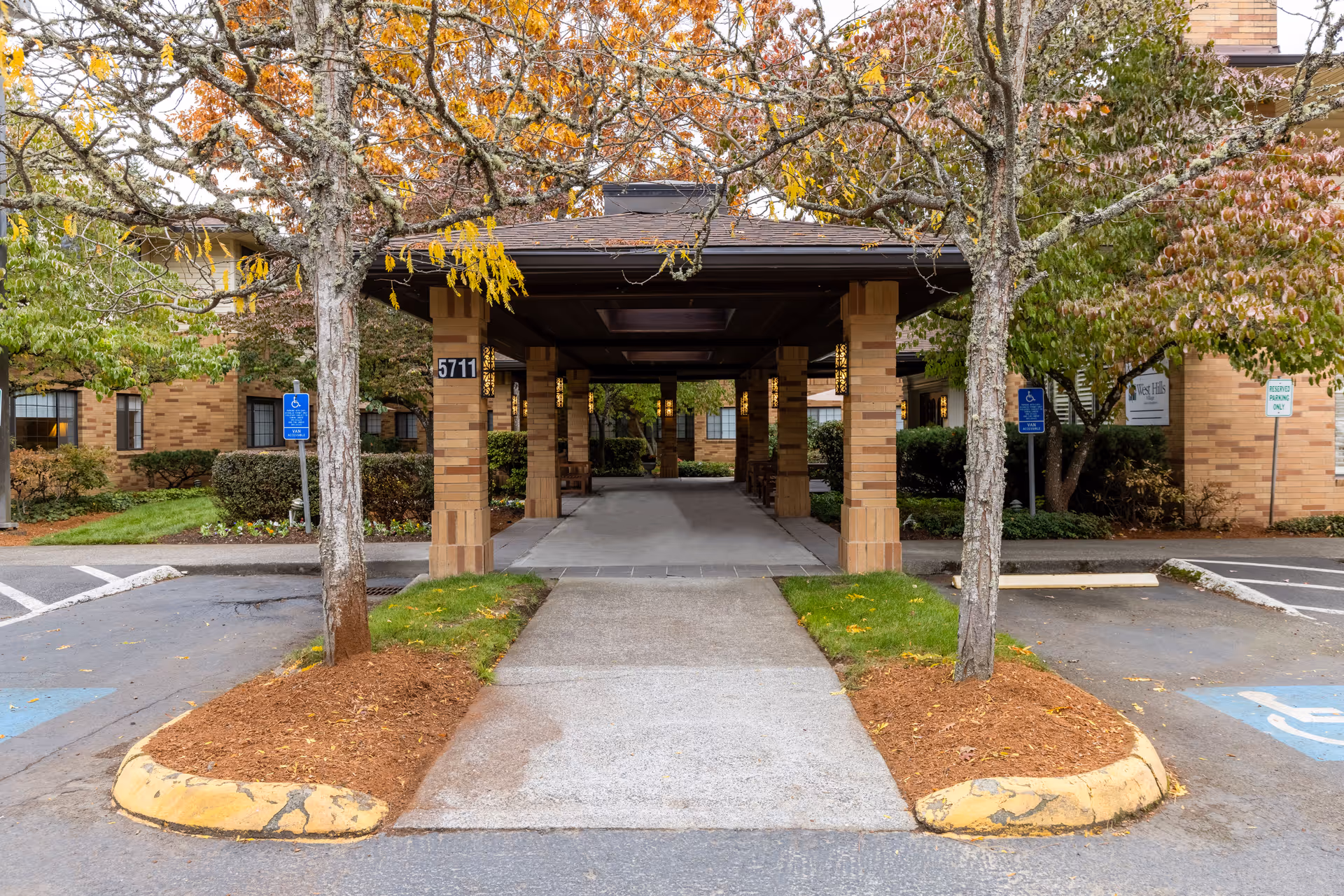 Covered porte-cochère entrance to a brick senior residence with columns, trees, and adjacent handicap parking spaces.