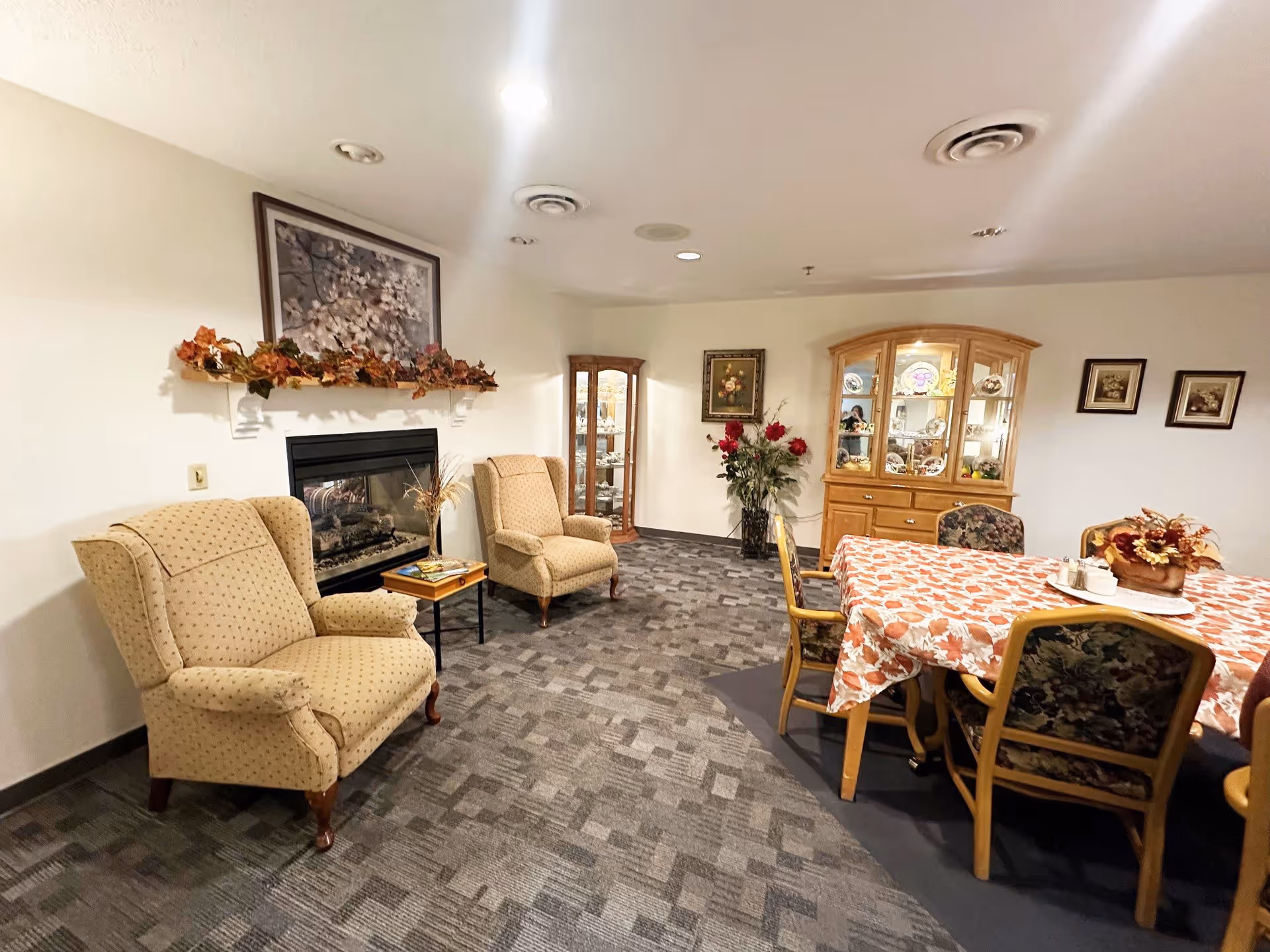 A cozy interior room in an assisted living facility featuring two beige upholstered armchairs near a gas fireplace decorated with autumn leaves and a floral painting above. To the right, there is a wooden dining table covered with a fall-themed tablecloth surrounded by chairs with floral upholstery. In the background, a wooden china cabinet displays decorative plates and other items, with framed artwork and a vase of red flowers on the wall.