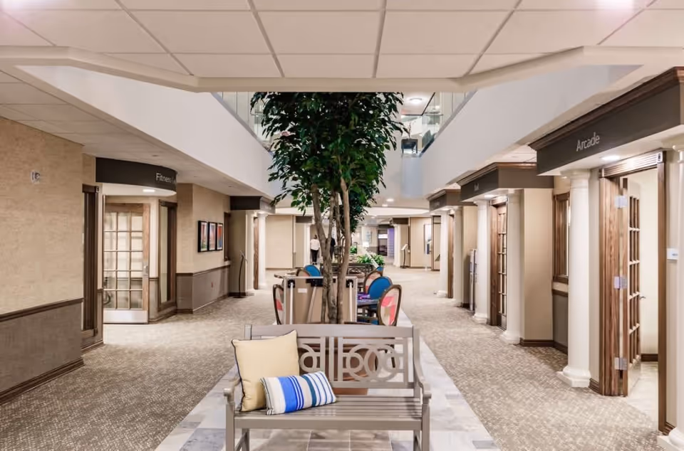 Interior hallway of Seaside Springs Retirement Community featuring a central seating area with a bench and cushions, a large indoor tree, and multiple doorways leading to different rooms including a fitness room and an arcade. The hallway has carpeted floors, beige walls with wooden trim, and a ceiling with recessed lighting.
