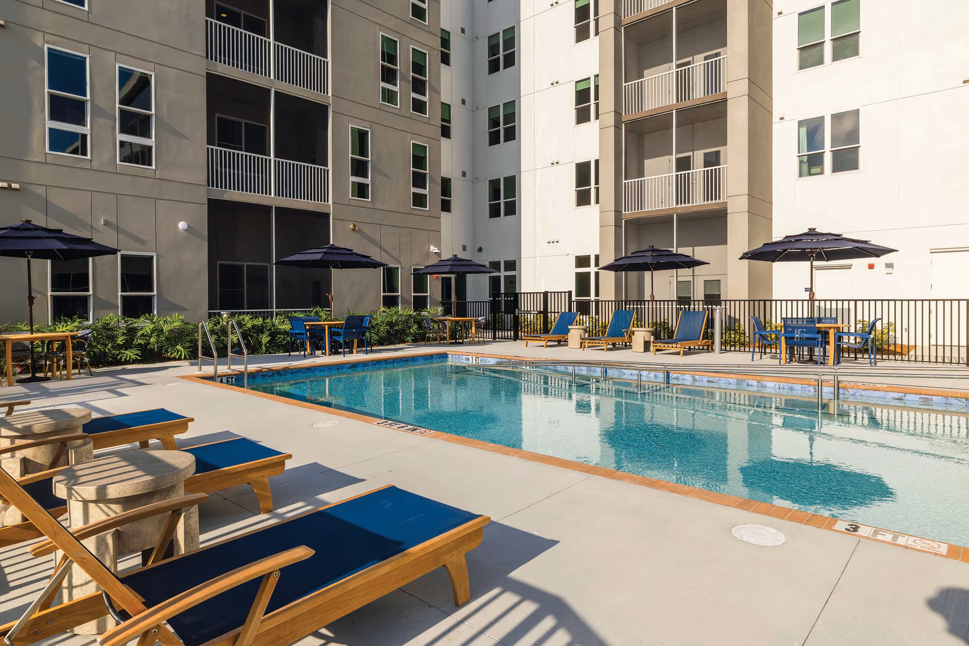 Outdoor swimming pool and lounge area in a multi-story residential courtyard with lounge chairs, tables, and umbrellas.