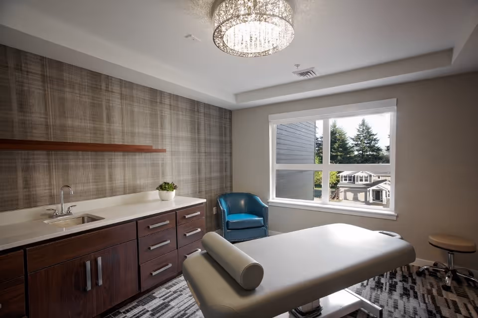 A bright treatment room with a massage/exam table, sink and wood cabinetry, a teal armchair and a large window overlooking neighboring houses.