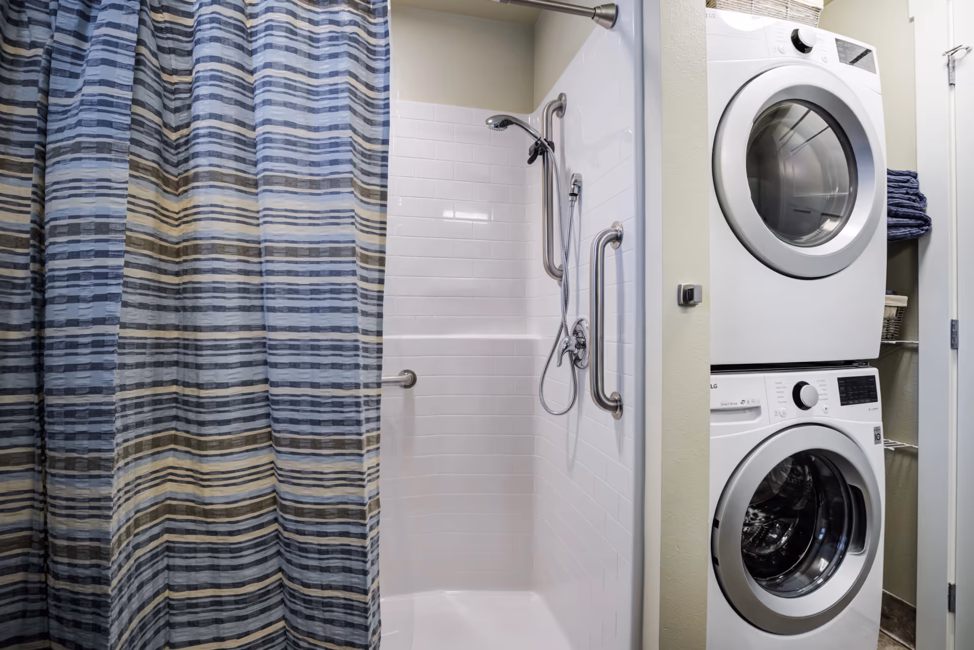 A bathroom shower area with white tiled walls and multiple stainless steel grab bars, a handheld showerhead, and a striped shower curtain in shades of blue, gray, and beige. Next to the shower is a stacked white washer and dryer unit with shelves holding folded towels and baskets.