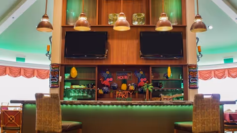 Interior view of a bar area with two flat-screen TVs mounted on a wooden wall. The bar counter has green lighting underneath and is decorated with colorful party-themed items including a sign that says 'Happy Hour' and 'Party Time'. There are two cushioned bar stools in front of the counter and tiki-style decorations on the sides. The background shows windows with pink valances and a bright, well-lit room.
