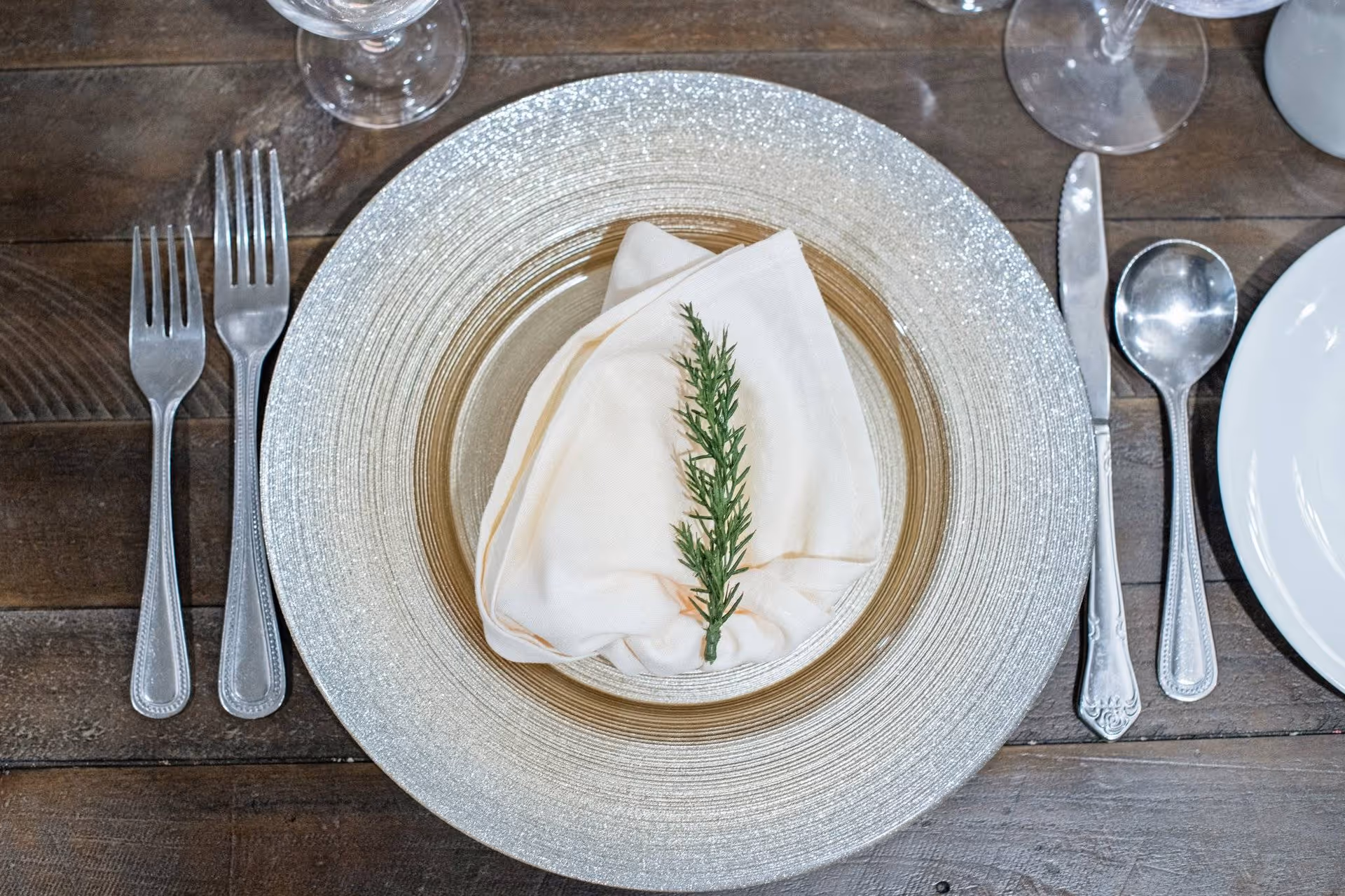 Close-up of a place setting with a silver charger plate, a folded napkin topped with a sprig of rosemary, and surrounding silverware on a wooden table.