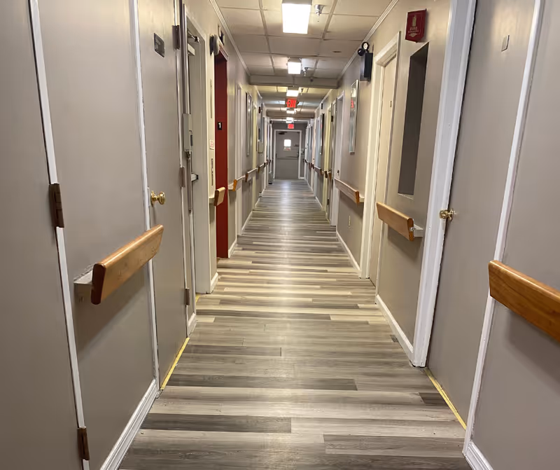 Long indoor hallway with multiple closed doors on both sides, handrails along the walls, and ceiling lights. The floor has wood-patterned vinyl or laminate flooring, and there are exit signs visible at the far end of the corridor.
