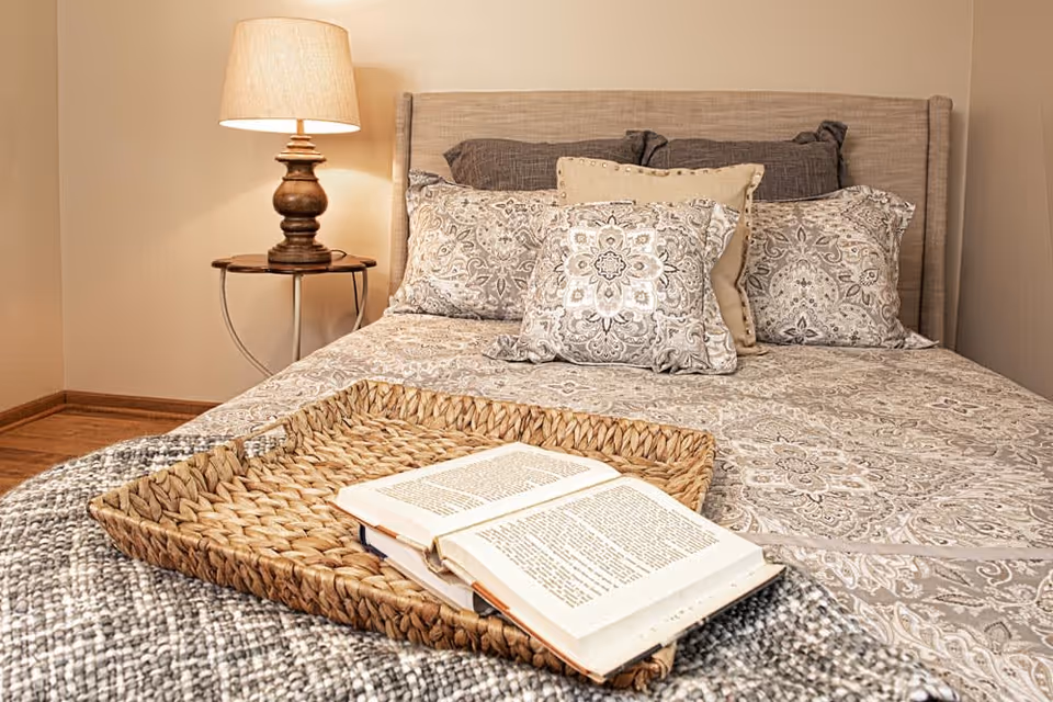 A neatly made bed with patterned pillows and a matching bedspread in neutral tones. A woven tray with an open book rests on the bed. A wooden side table with a decorative lamp is positioned next to the bed against a beige wall.