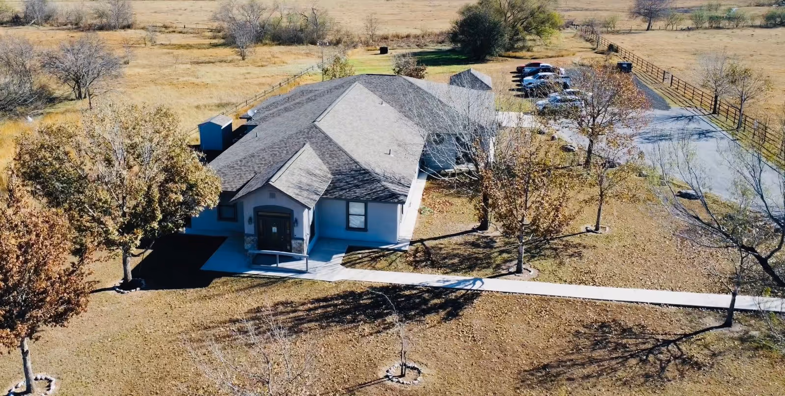 Aerial view of a single-story building with a dark shingled roof surrounded by a grassy area with several trees and a paved walkway leading to the entrance. There is a parking area with several cars visible to the right of the building, and the surrounding landscape is open with sparse trees and fencing.