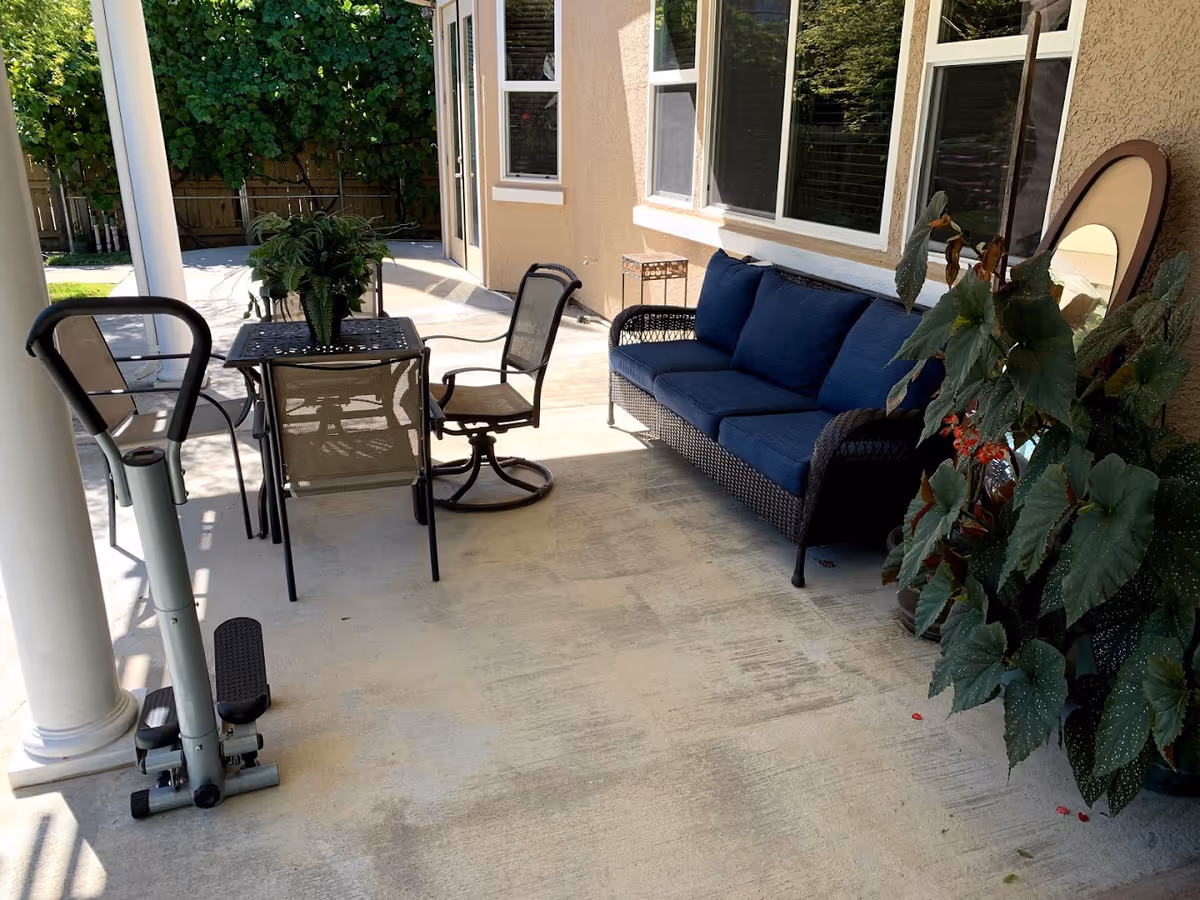 Outdoor patio area with a black metal table and four chairs, a blue cushioned wicker sofa, a large potted plant, and a small exercise stepper machine near a white column. The patio is adjacent to a beige building with windows and a door, surrounded by greenery and a wooden fence.