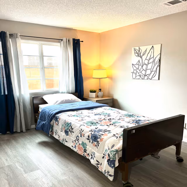 A tidy resident bedroom featuring floral bedding, a bedside table with lamp, and blue curtains.