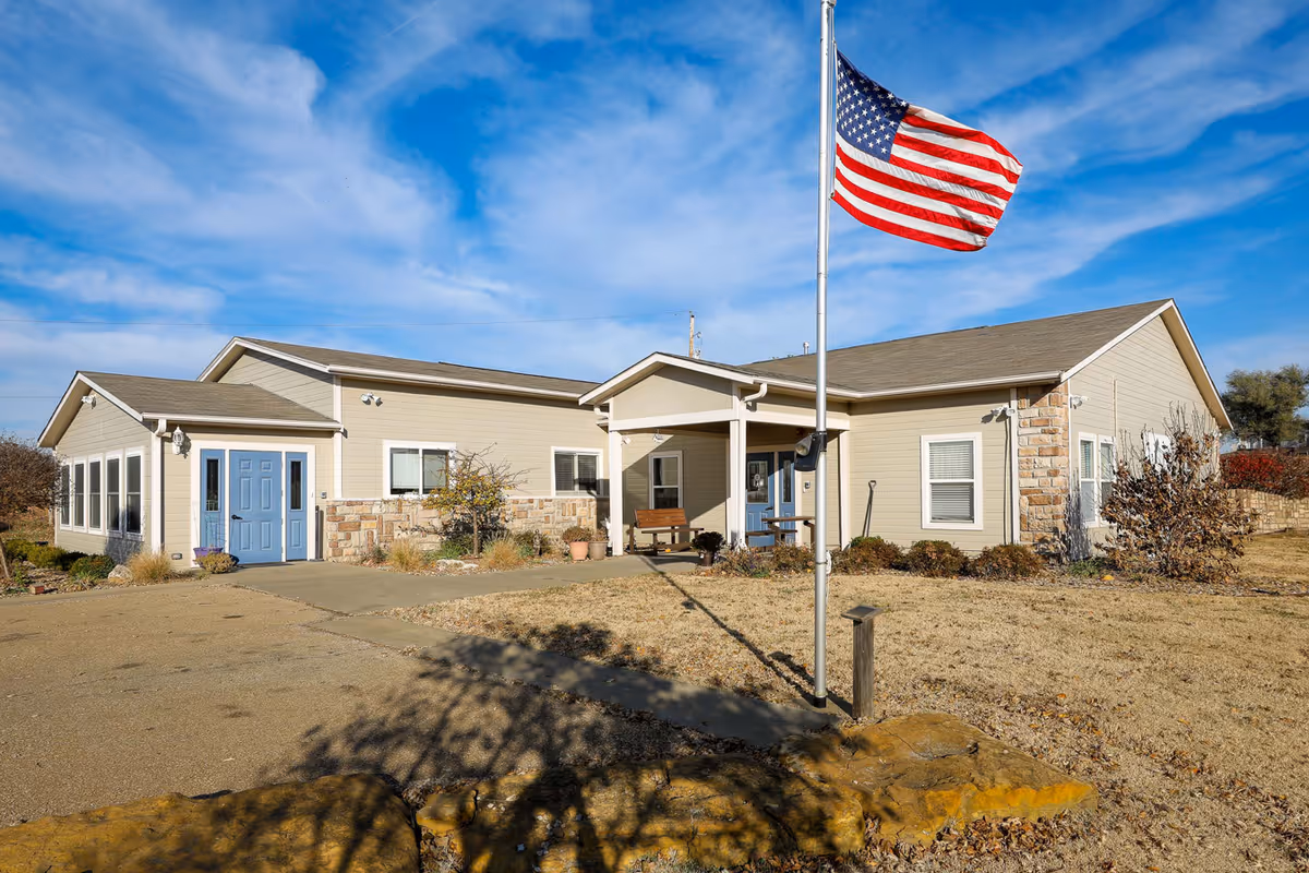 Single-story beige building with blue doors, a covered front entrance, landscaped grounds, and an American flag on a flagpole.