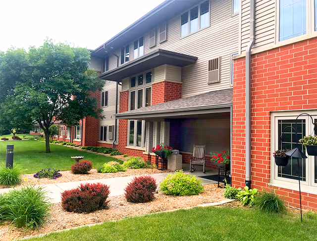 Front exterior of a multi-story brick and siding senior living building with a landscaped walkway, porch seating, and potted plants.