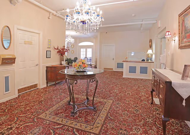 A spacious, well-lit reception area with ornate red patterned carpet, a round glass table with a floral centerpiece in the middle, chandeliers hanging from the ceiling, and classic wooden furniture along the walls. There is a reception desk on the right side and double doors at the far end of the room.