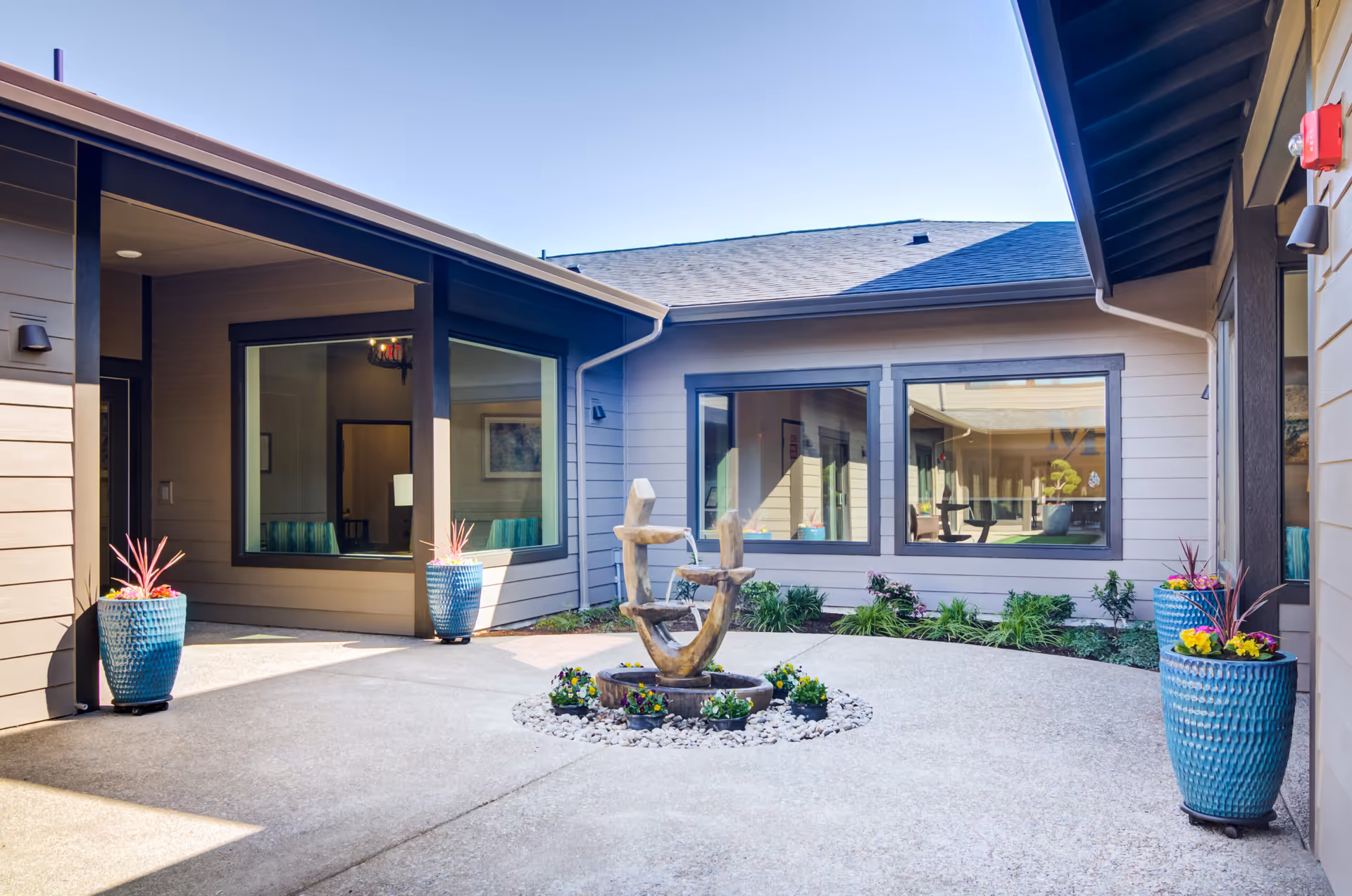 Outdoor courtyard area at Miracle Heights featuring a modern abstract water fountain surrounded by small flower pots and decorative blue planters with colorful plants. The courtyard is enclosed by building walls with large windows showing interior rooms and a clear blue sky overhead.