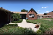 Brick single-story courtyard with a concrete walkway, potted plants, grassy areas, and a blue sky.