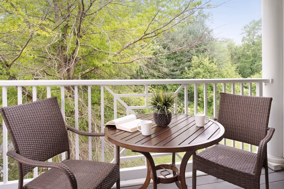 A balcony with two brown wicker chairs and a round wooden table. On the table are two white mugs, an open book, and a small potted plant. The balcony overlooks green trees and foliage.