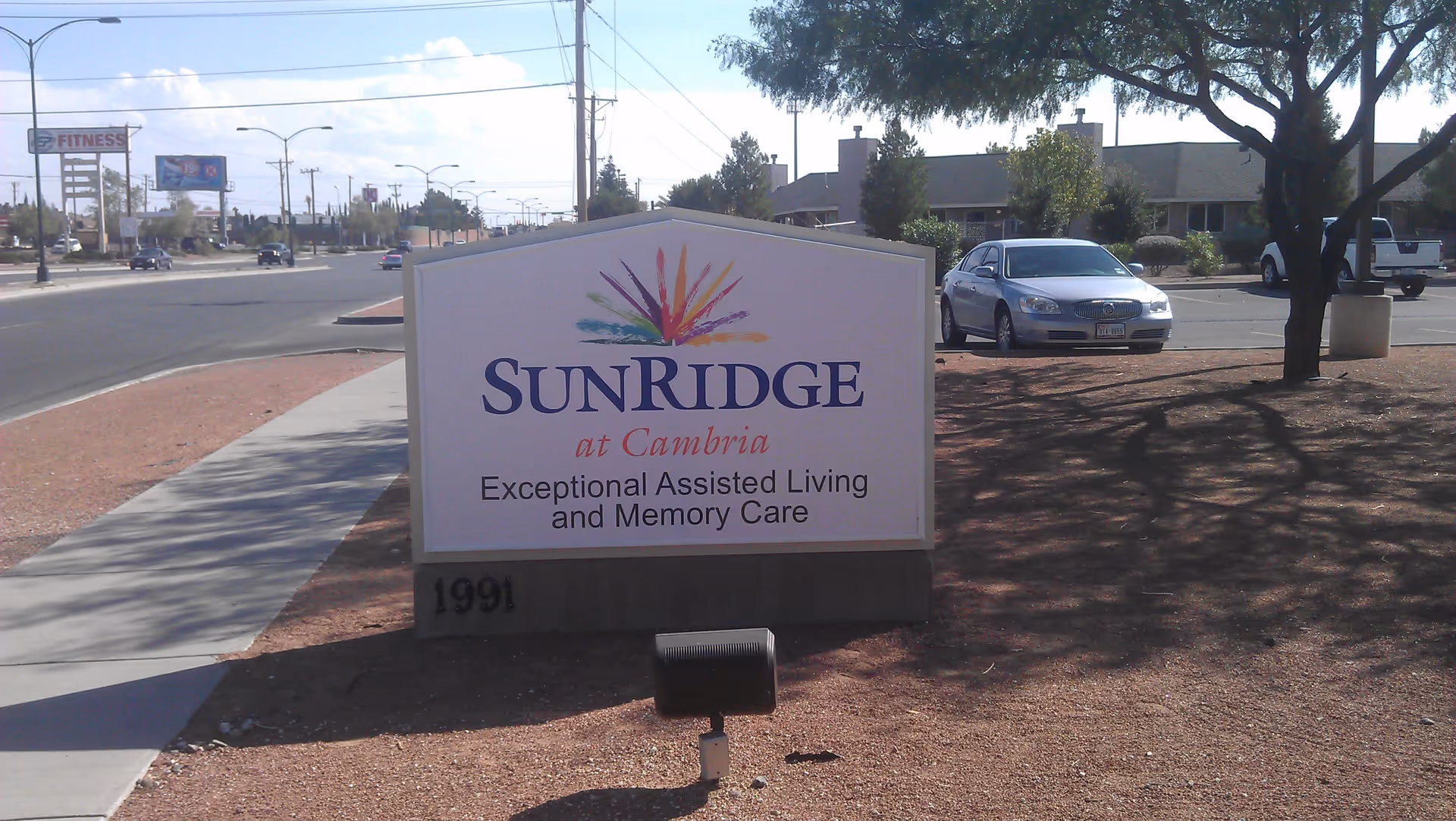 Outdoor view of a sign for SunRidge at Cambria, an assisted living and memory care facility, located near a street with cars and a parking lot with trees and a silver car.