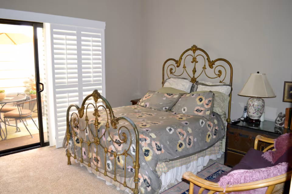 A bedroom with an ornate brass bed dressed in floral bedding, a sliding glass door with shutters, and a nightstand with a lamp.
