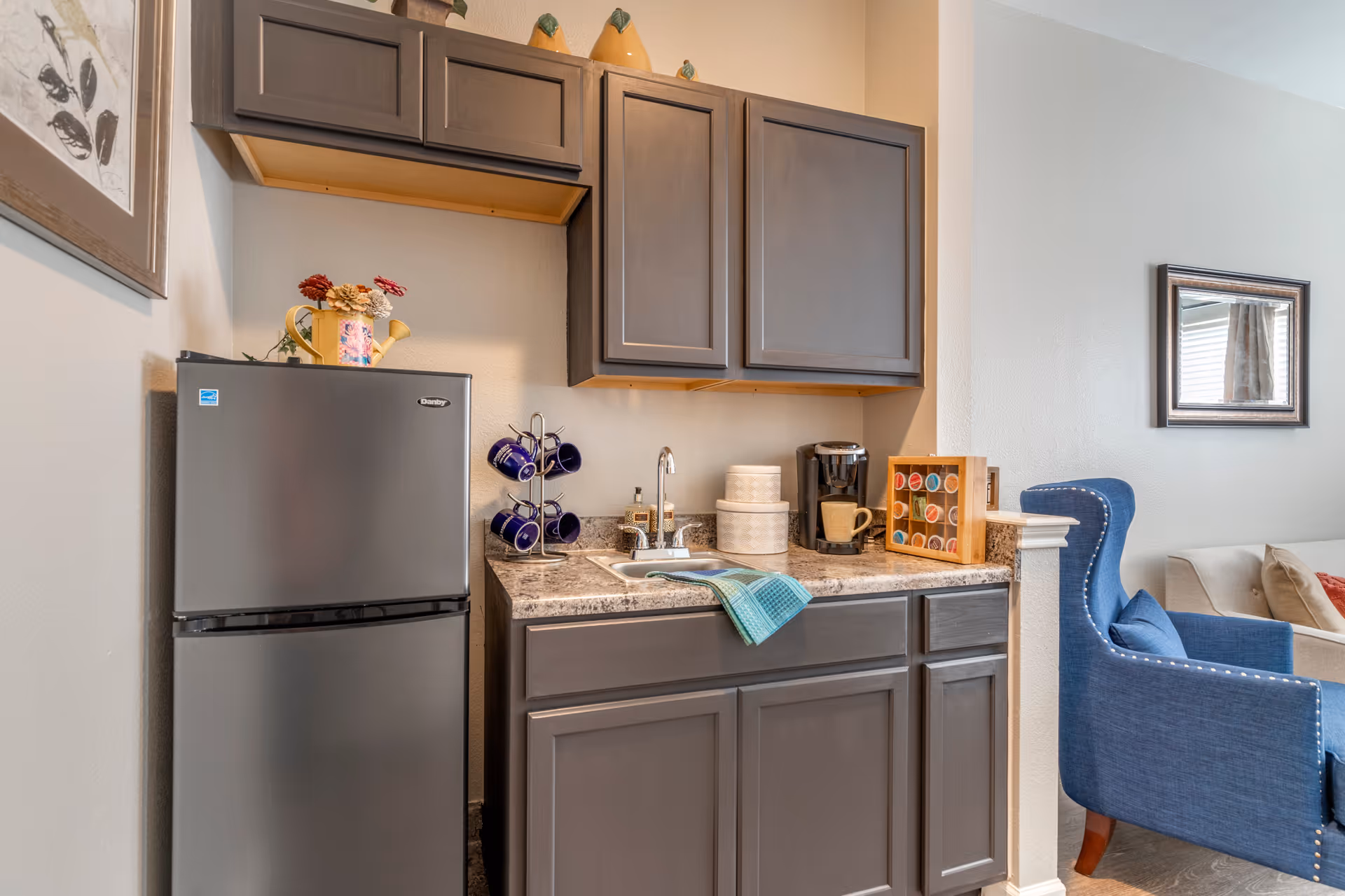 Small kitchenette area with gray cabinets, a stainless steel mini refrigerator, a sink with a blue dish towel, a coffee maker, a mug holder with blue mugs, and a wooden box holding coffee pods. Adjacent to the kitchenette is a blue upholstered armchair and a beige sofa with pillows, with a framed mirror on the wall.