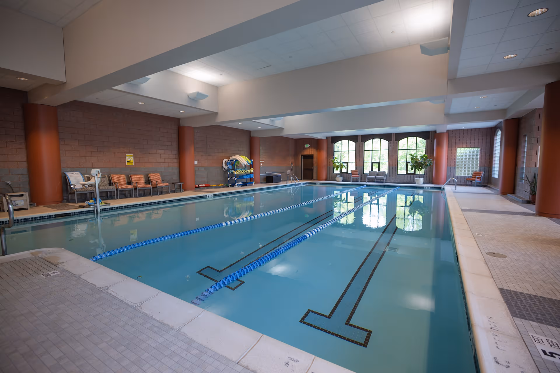 Indoor swimming pool with two swimming lanes, surrounded by tiled flooring and brick walls. There are chairs and pool equipment along the walls, large windows letting in natural light, and potted plants near the windows.