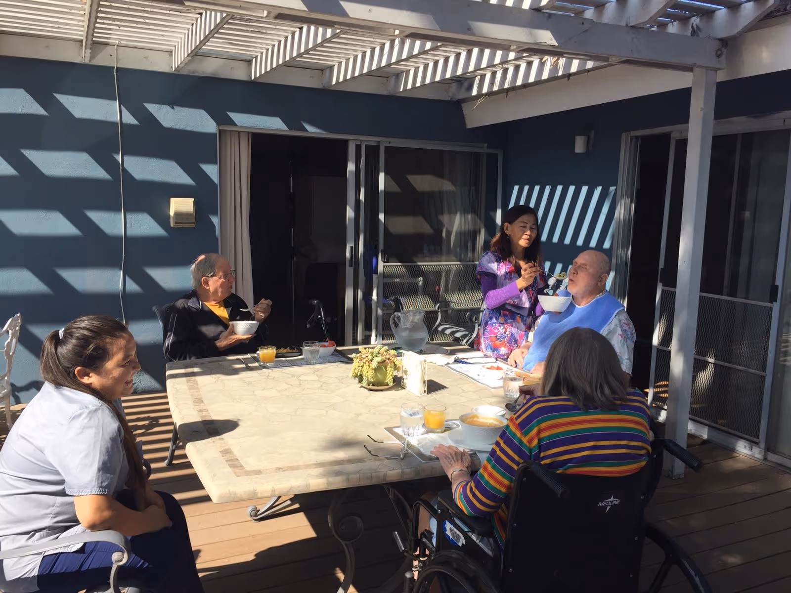 Four elderly individuals and a caregiver are gathered around a large outdoor table on a patio with a pergola overhead. One elderly man is being fed by the caregiver, while others are seated and eating or conversing. The setting is sunny with shadows cast by the pergola, and the background shows sliding glass doors leading inside.