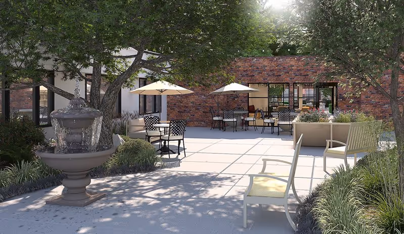 Outdoor patio area with several tables and chairs under umbrellas, surrounded by trees and greenery. A water fountain is visible on the left side, and a brick building with large windows is in the background.