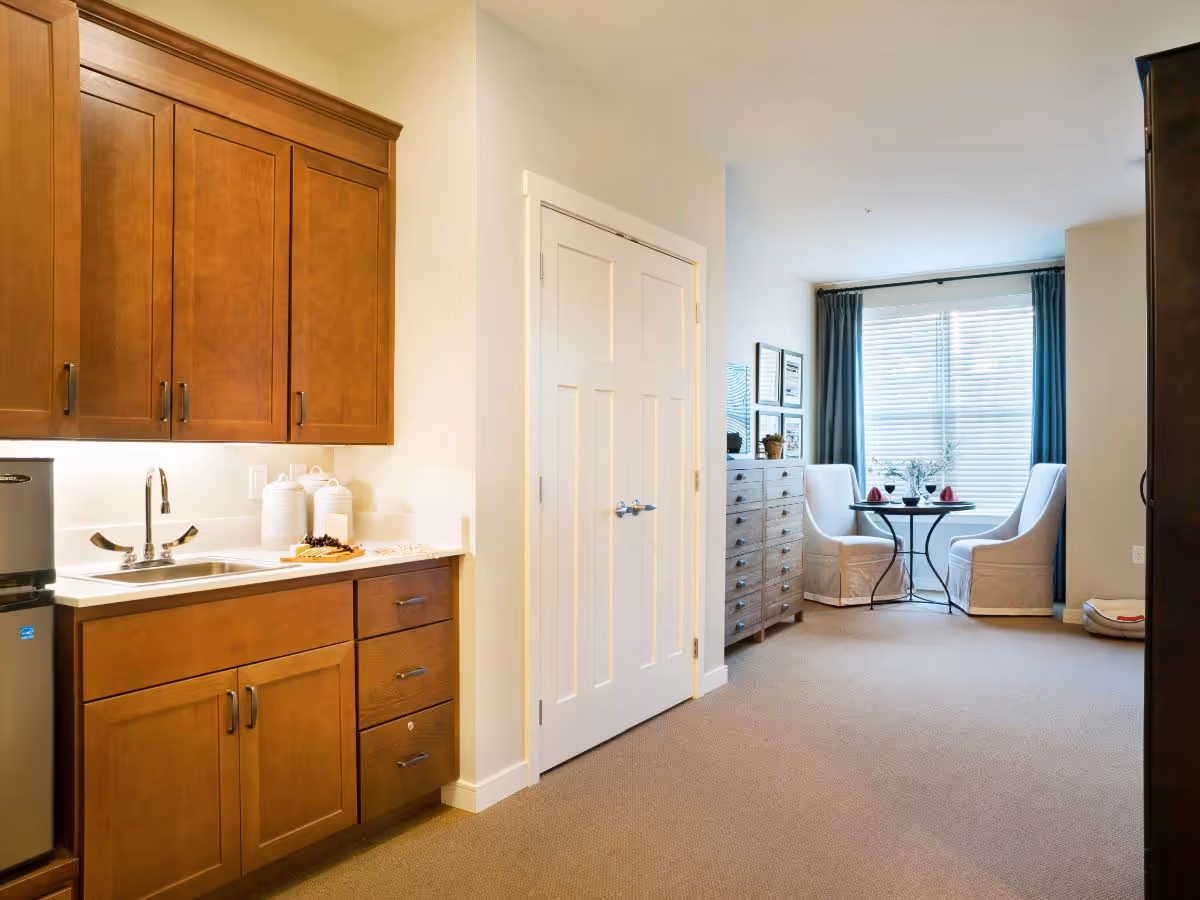Interior view of a senior living facility room with a small kitchenette featuring wooden cabinets, a sink, and a mini refrigerator on the left. In the background, there is a cozy dining area with a round table set for two, two upholstered chairs, a chest of drawers, and a large window with blue curtains letting in natural light.