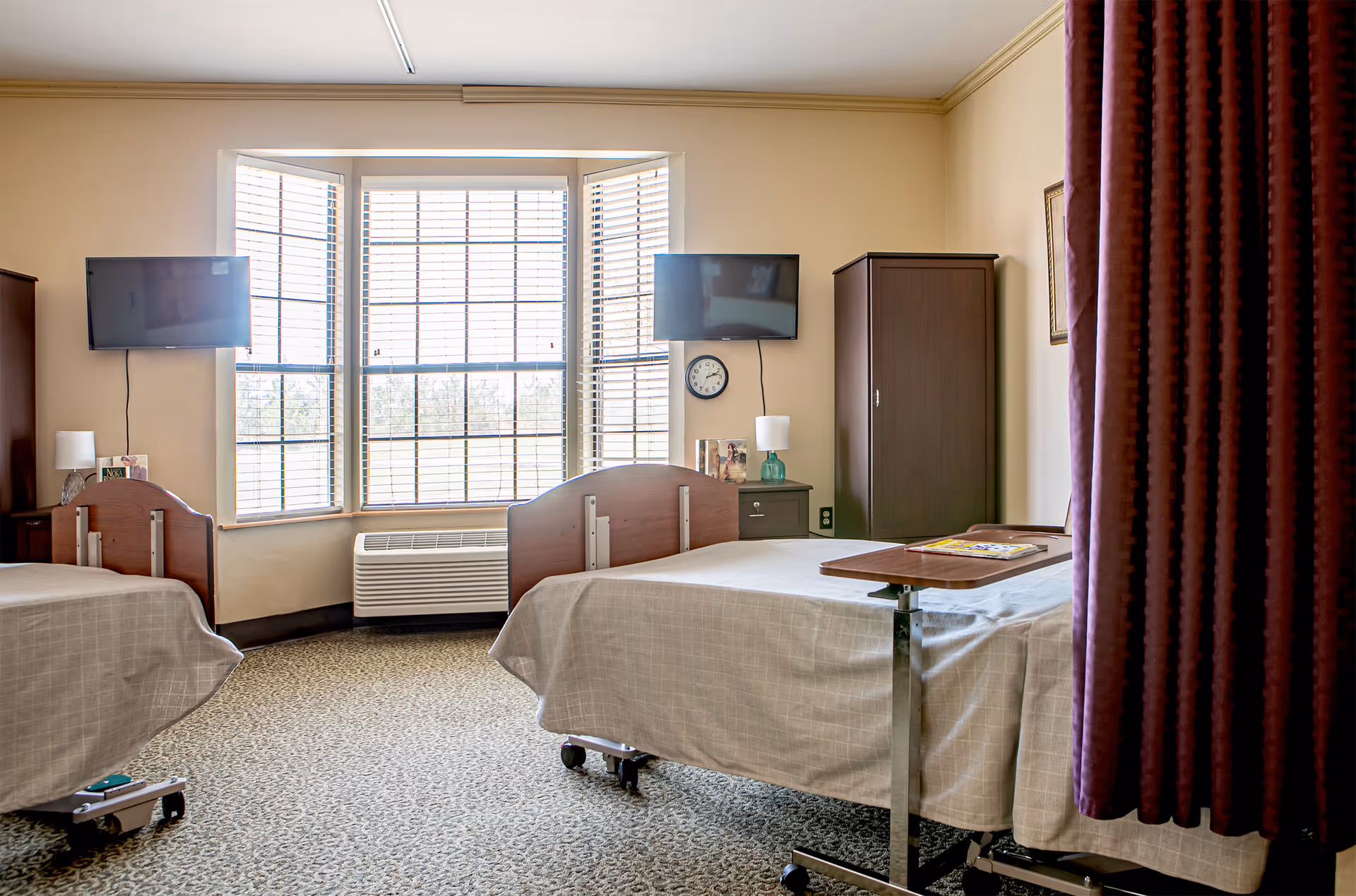 A bright room with two hospital-style beds covered in beige bedspreads, each with a mounted TV on the wall above. Between the beds is a large window with white blinds, a small nightstand with a lamp, and a clock on the wall. There is a dark wooden wardrobe and a curtain partially visible on the right side.