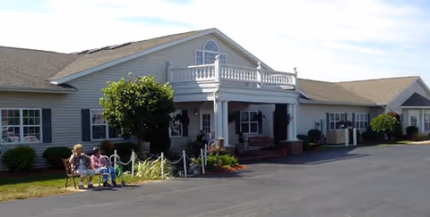 Exterior view of a single-story assisted living community building with white siding, a covered entrance with columns, and a small balcony above. There are shrubs and small trees around the building, and three people sitting on a bench near the entrance. The sky is clear and the driveway is paved.