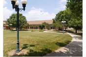 A single-story brick building fronts a grassy courtyard with lamp posts and a curved walkway under a partly cloudy sky.