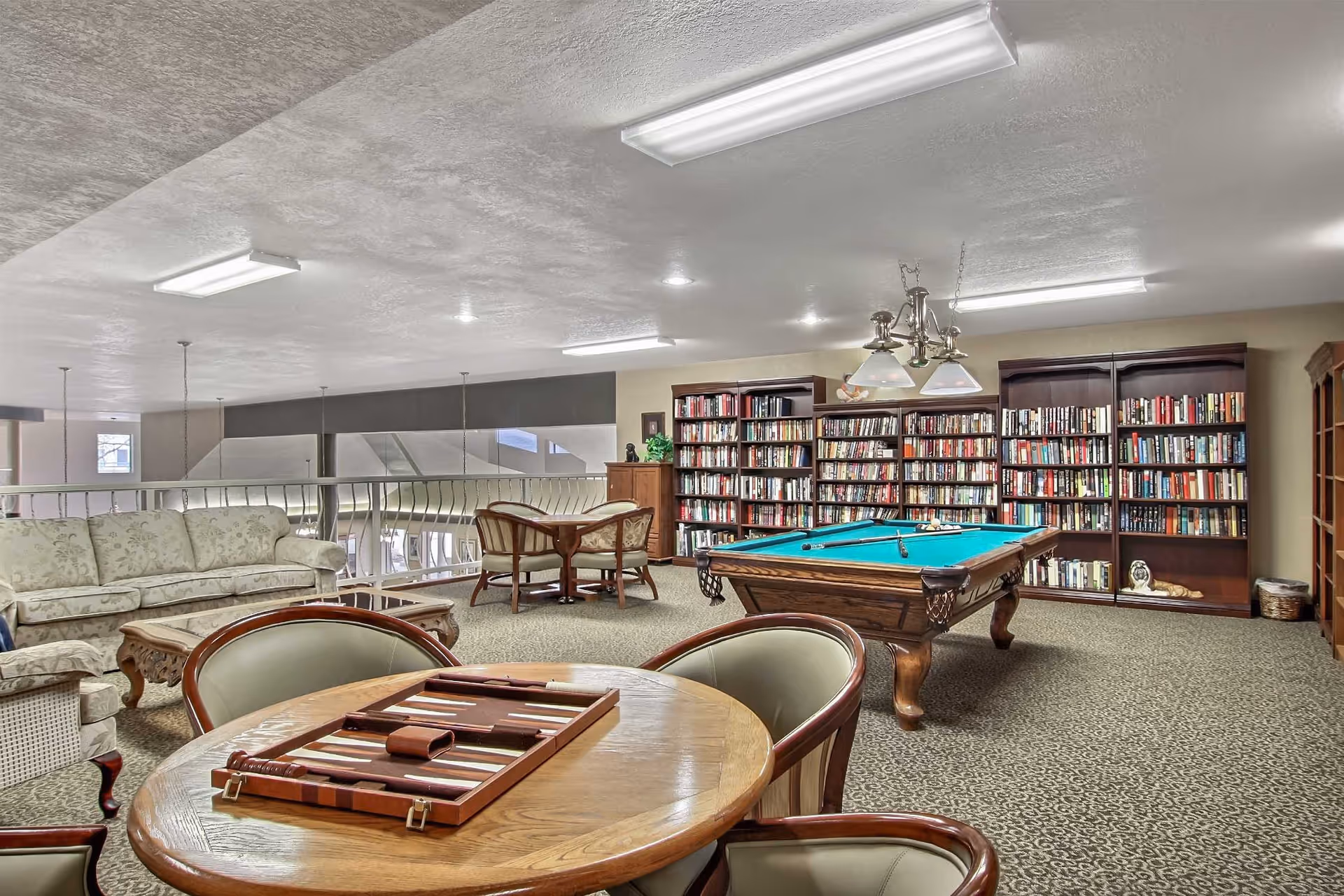 A senior living common room with a pool table, bookshelves, sofas, and card tables.