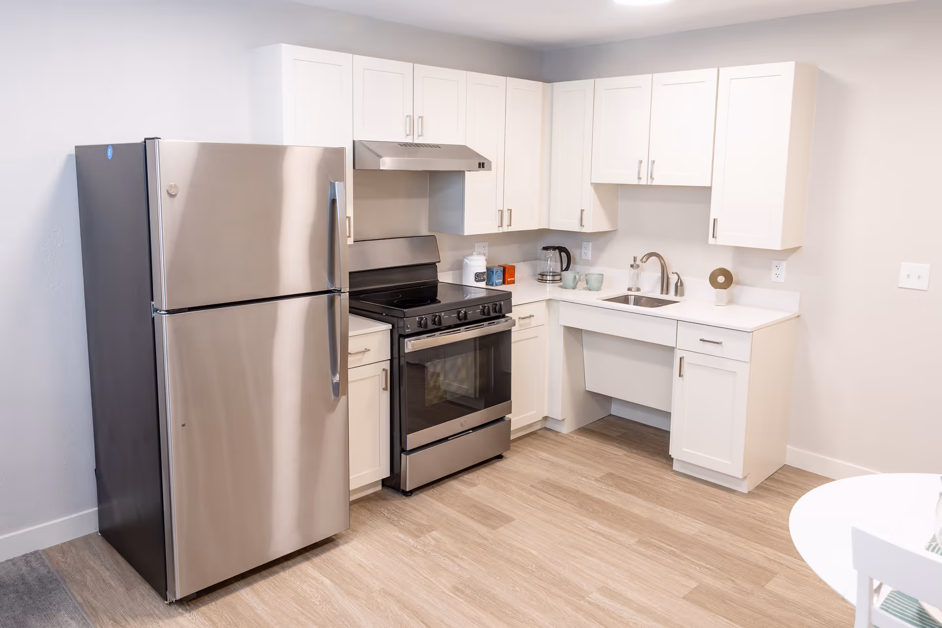 A modern kitchen with white cabinets, a stainless steel refrigerator, a stainless steel stove with an overhead vent, a small sink with a faucet, and a few kitchen items on the countertop including a coffee pot, mugs, and containers.