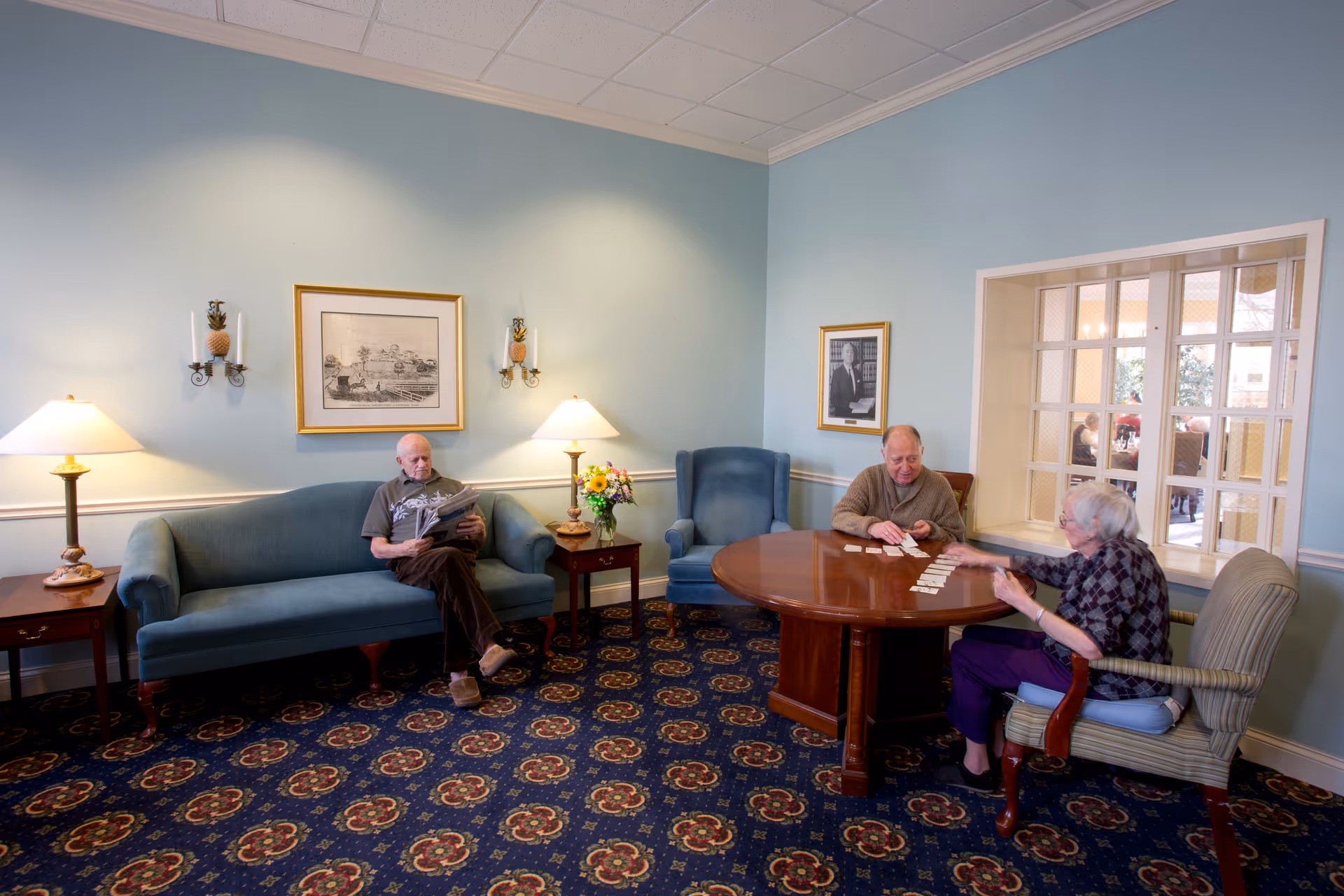 Three elderly residents in a comfortable common room — one reading on a sofa and two playing cards at a round table.
