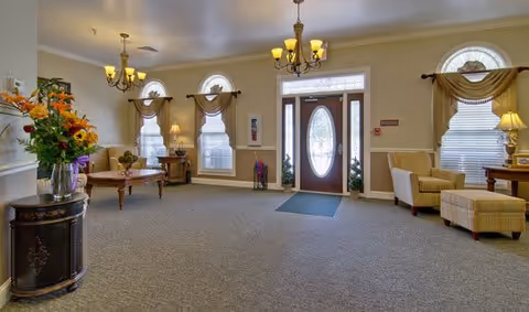 Spacious senior living facility lobby with front door, seating areas, chandeliers, and decorative arched windows.