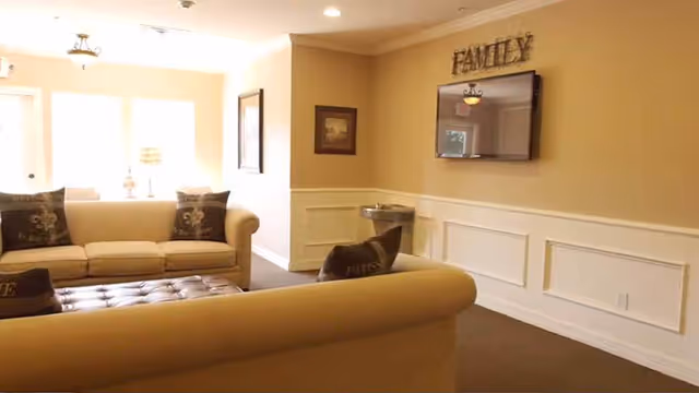 A cozy living room area with beige walls and white wainscoting. There are two beige sofas with dark decorative pillows facing each other, and a dark tufted ottoman in between. On the wall above a mounted flat-screen TV, the word 'FAMILY' is displayed in decorative letters. A water fountain is visible in the corner, and framed pictures hang on the walls. Natural light comes through a window in the background.