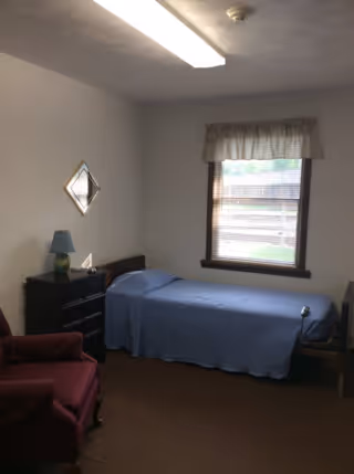 A small bedroom with a single bed covered in a blue bedspread, a wooden nightstand with a blue lamp, a red upholstered armchair, a window with blinds and a valance, and a diamond-shaped mirror on the wall.