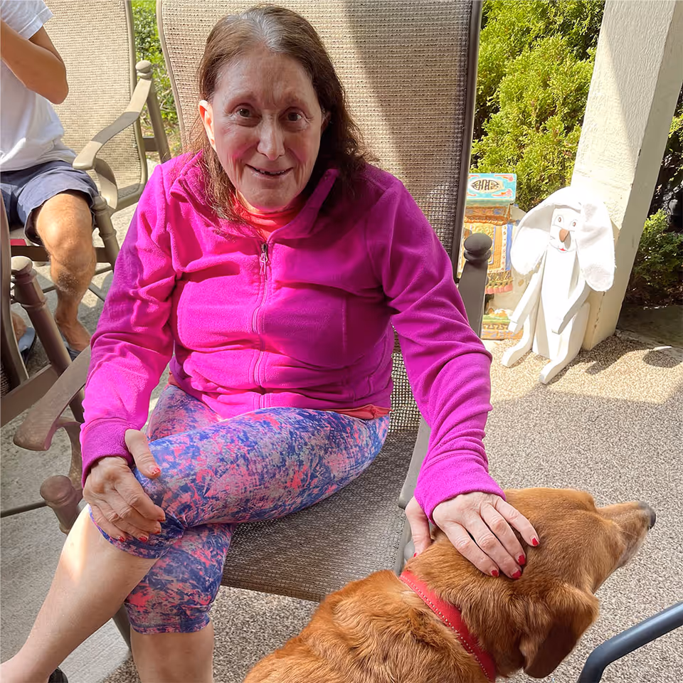 An elderly woman wearing a bright pink jacket and colorful leggings is sitting on a patio chair, smiling and petting a brown dog with a red collar. There is a person sitting next to her wearing a white shirt and dark shorts. In the background, there is greenery and a white decorative figure resembling a dog.