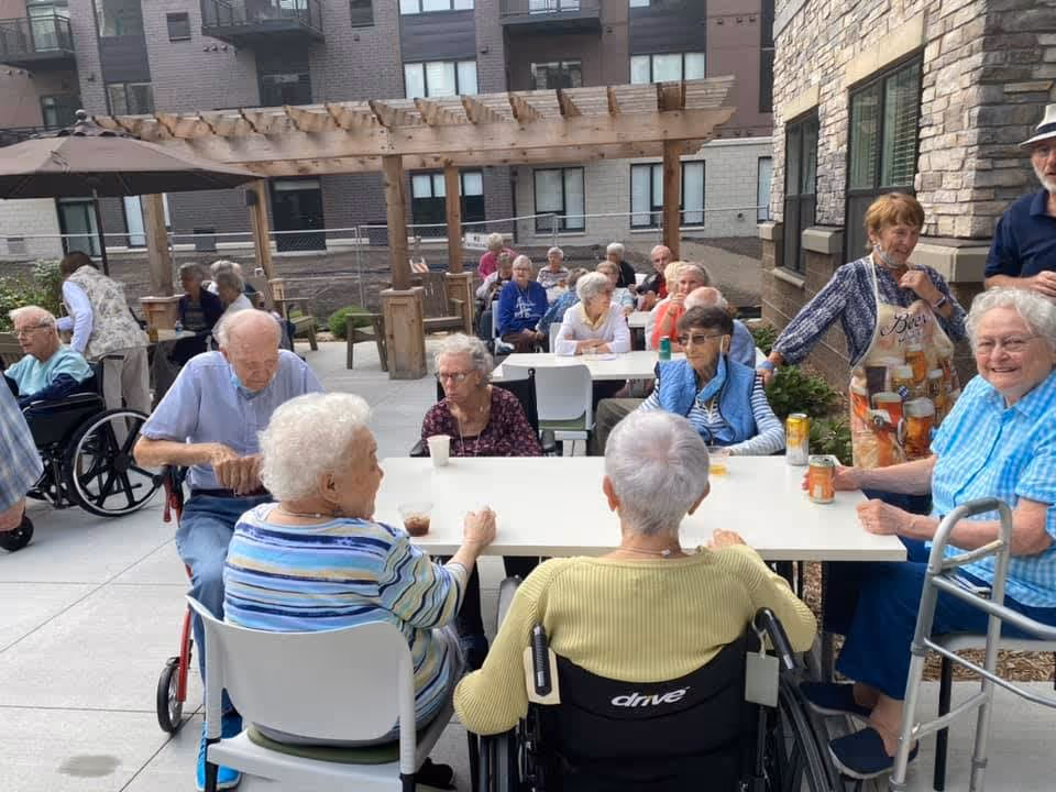 A group of elderly people sitting and socializing at tables in an outdoor patio area of a senior living facility. Some are in wheelchairs, and others are seated on chairs. The patio has a pergola, umbrellas, and is surrounded by the building's exterior walls. One woman is standing and wearing an apron, engaging with the group.