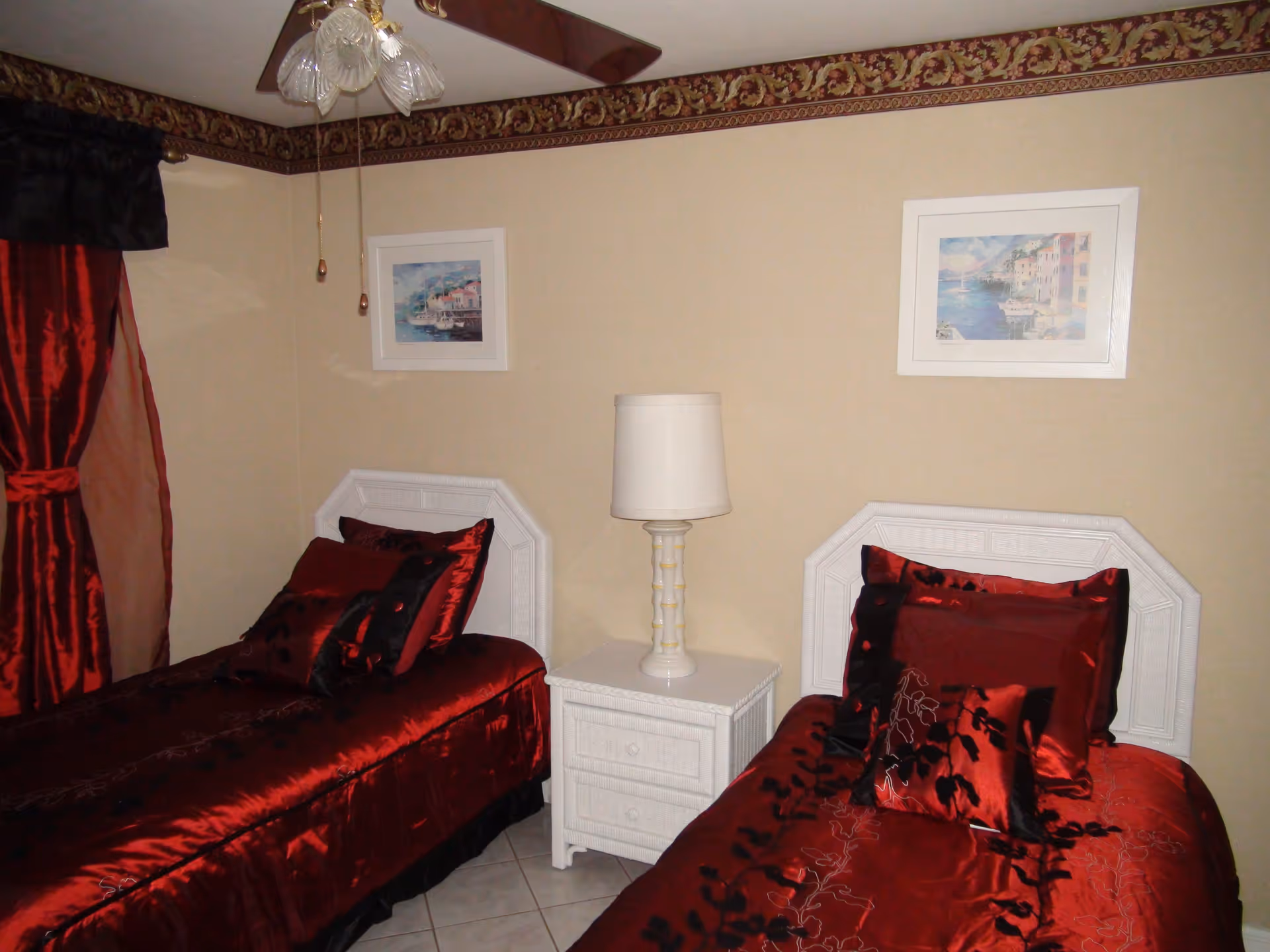 A bedroom with two twin beds featuring white headboards and red satin bedding with black floral patterns. Between the beds is a white nightstand with a white lamp. The walls are beige with a decorative border near the ceiling, and two framed pictures hang above each bed. Red and black curtains cover a window on the left side.